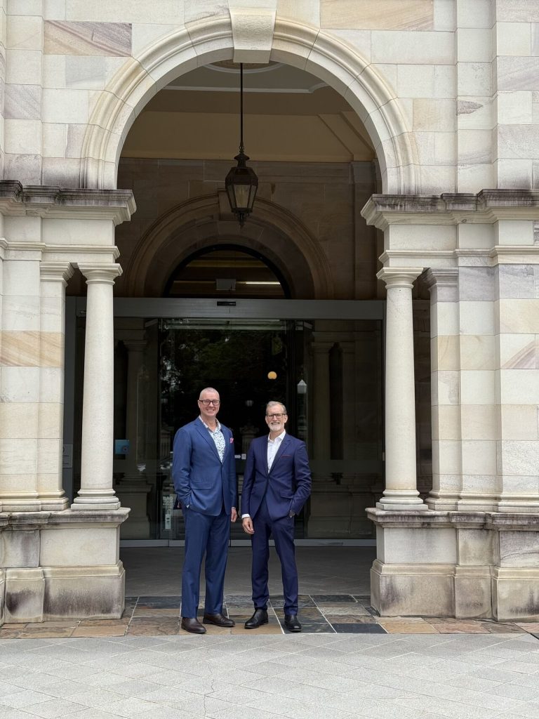 Two men standing in archway of an old building wearing navy blue suits. 